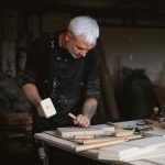 Serious male woodworker in apron using mallet and chisel to carve wooden board at table with abundance of instruments in carpentry